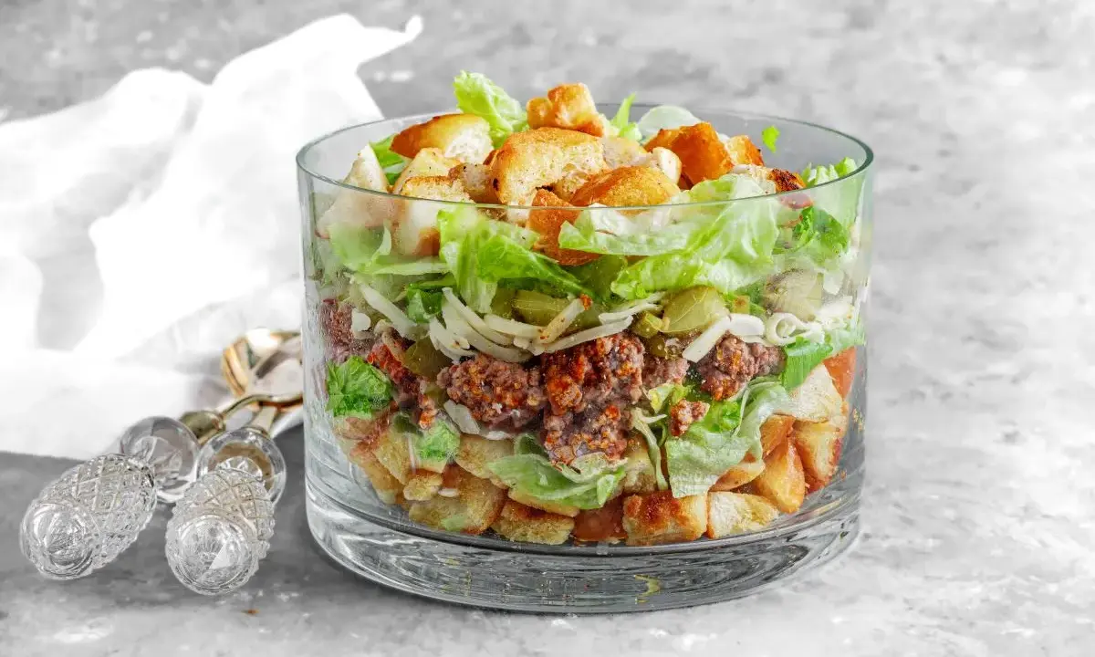 Warstwowa sałatka big mac w szklanym naczyniu, z mielonym mięsem, sałatą, serem, ogórkiem i grzankami.