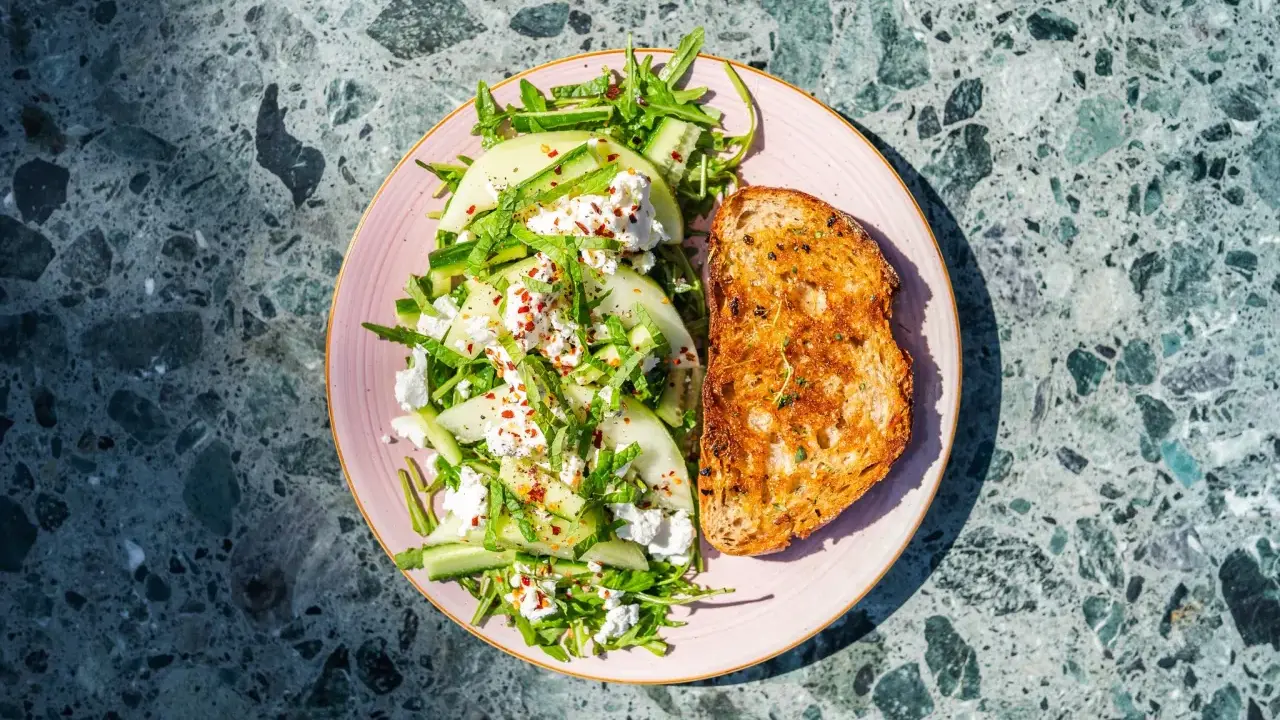 Orzeźwiająca sałatka z melonem, serem feta, ogórkiem i rukolą, posypana płatkami chili. Obok tost z przyprawami.