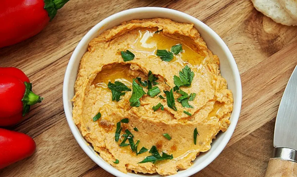 Pyszny hummus z suszonymi pomidorami, polany oliwą i posypany natką pietruszki, obok czerwonej papryki i pieczywa.