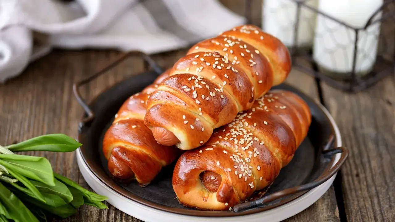 Złociste parówki w cieście drożdżowym, posypane sezamem, kuszą na rustykalnym talerzu.