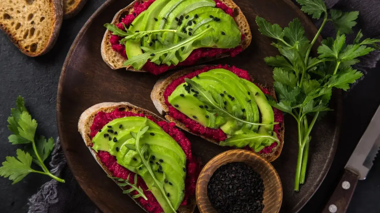 Trzy kanapki z awokado na ciemnym pieczywie, posypane czarnym sezamem i rukolą, na drewnianej desce.