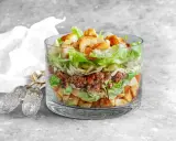 Warstwowa sałatka big mac w szklanym naczyniu, z mielonym mięsem, sałatą, serem, ogórkiem i grzankami.