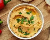Pyszny hummus z suszonymi pomidorami, polany oliwą i posypany natką pietruszki, obok czerwonej papryki i pieczywa.