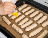 Ręka pędzlem smaruje parówki w cieście francuskim ułożone na blasze.