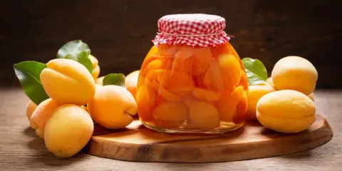 Słoik pełen soczystych, domowych moreli w syropie, otoczony świeżymi owocami i liśćmi.