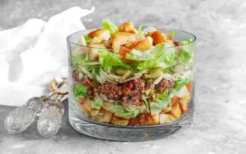 Warstwowa sałatka big mac w szklanym naczyniu, z mielonym mięsem, sałatą, serem, ogórkiem i grzankami.