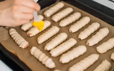 Ręka pędzlem smaruje parówki w cieście francuskim ułożone na blasze.