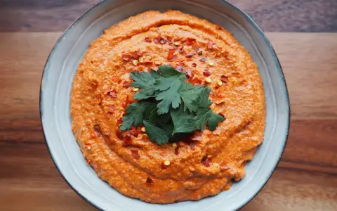 Aksamitna pasta z papryki, posypana płatkami chili i ozdobiona gałązką pietruszki, w niebieskiej misce na drewnianym tle.