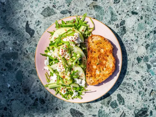 Orzeźwiająca sałatka z melonem, serem feta, ogórkiem i rukolą, posypana płatkami chili. Obok tost z przyprawami.