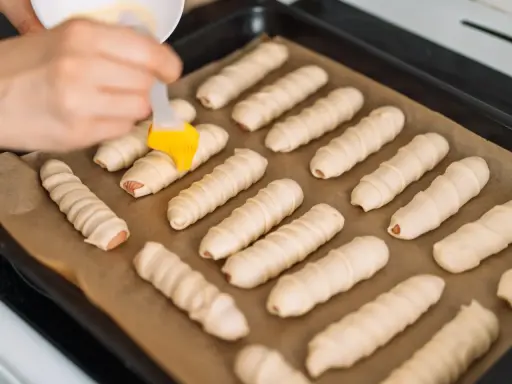 Ręka pędzlem smaruje parówki w cieście francuskim ułożone na blasze.