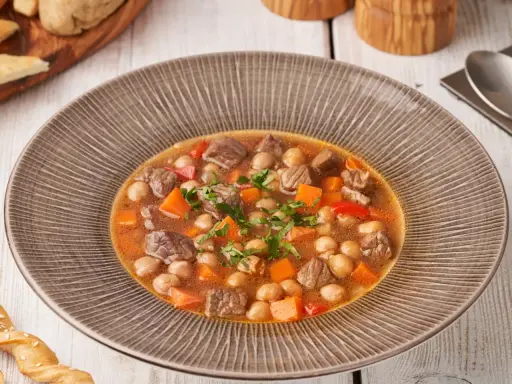 Gęsty gulasz z ciecierzycy, kawałków mięsa i marchewki, posypany natką pietruszki, podany w misce.