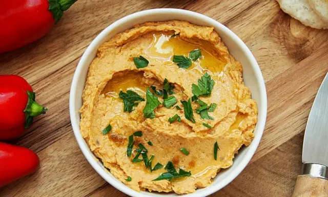 Pyszny hummus z suszonymi pomidorami, polany oliwą i posypany natką pietruszki, obok czerwonej papryki i pieczywa.