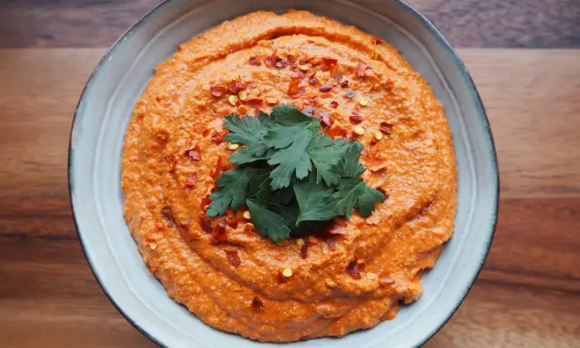 Aksamitna pasta z papryki, posypana płatkami chili i ozdobiona gałązką pietruszki, w niebieskiej misce na drewnianym tle.