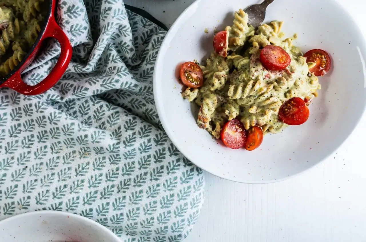 Pyszna pasta z awokado w kremowym sosie, udekorowana połówkami pomidorków koktajlowych, podana na białym talerzu obok czerwonego garnka.