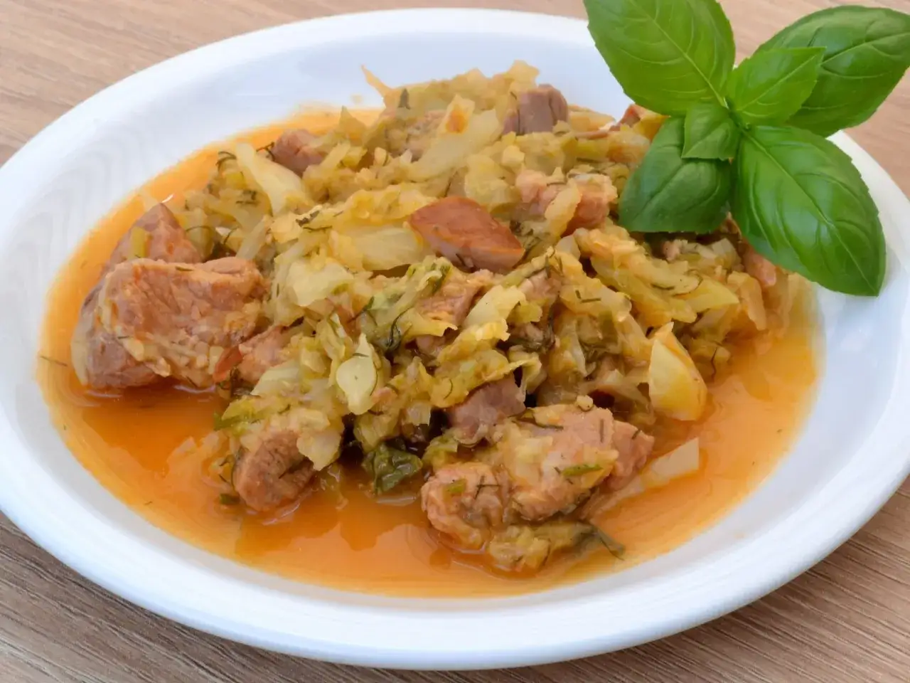 Pyszny bigos ze słodkiej kapusty z kawałkami mięsa i kiełbasy, udekorowany gałązką bazylii.