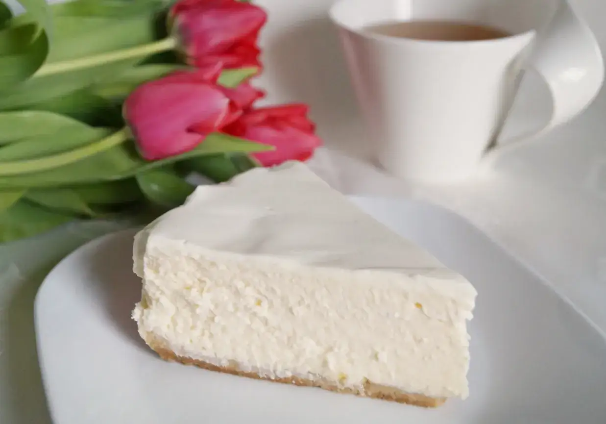 Pyszny sernik z mascarpone na białym talerzyku, obok bukiet różowych tulipanów i filiżanka herbaty.
