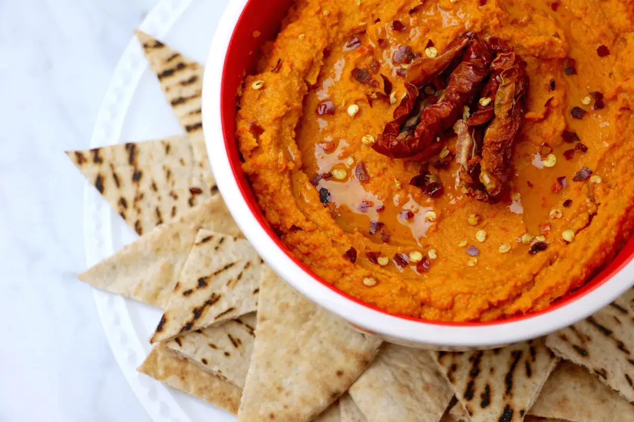 Pomarańczowy hummus z suszonymi pomidorami i płatkami chili, podany z grillowanymi kawałkami pity.